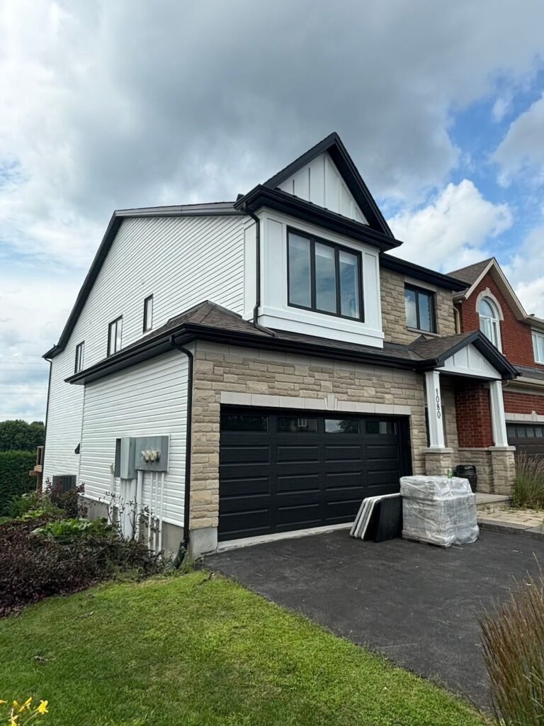 A modern two-story house with white siding, stone accents, and a black double garage door. Several large packages for exterior painting and black panels are stacked in the driveway. Lush green lawn and cloudy sky are visible.